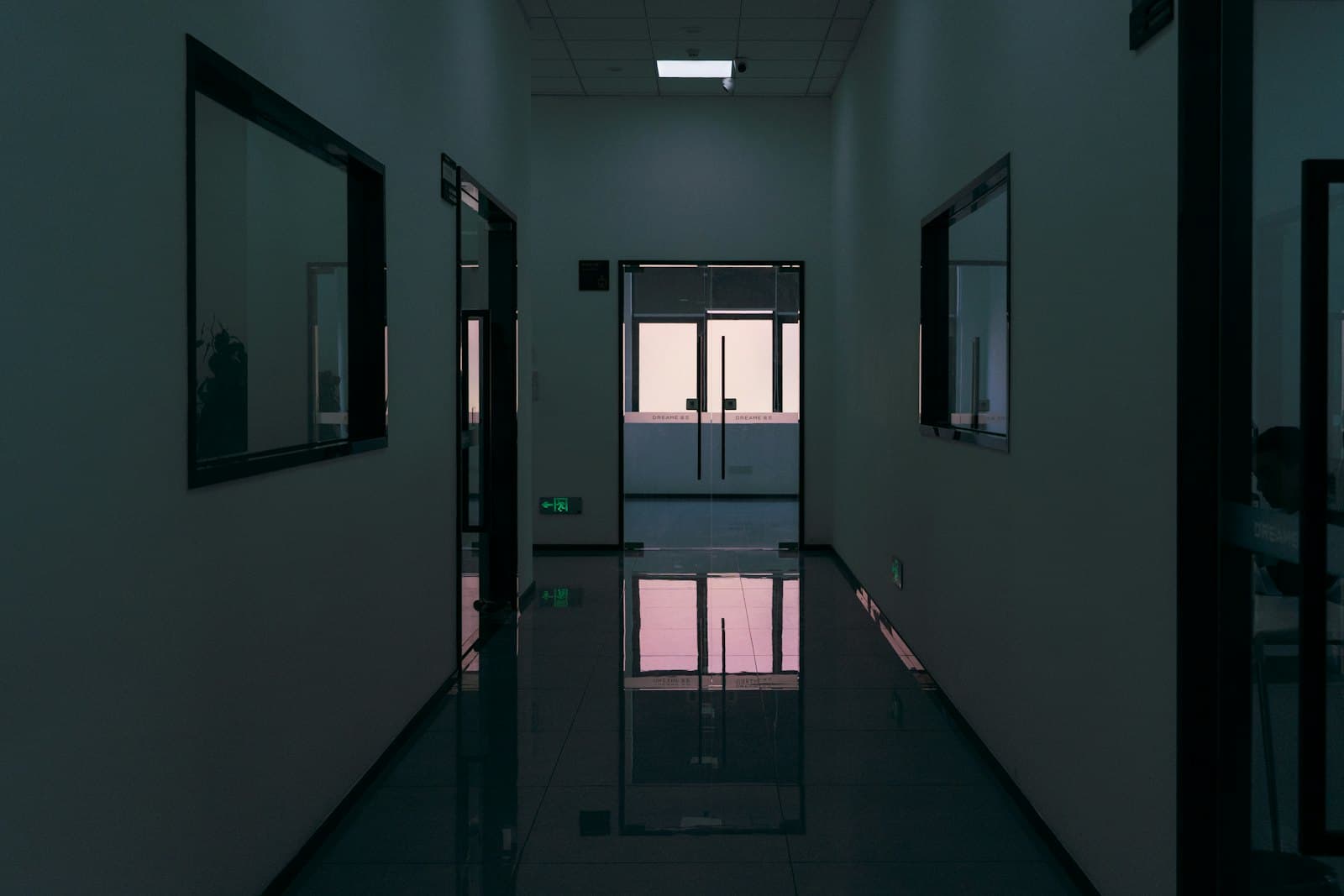 The corridor of an office building in the dark, emergency light on and the floor mirroring it.